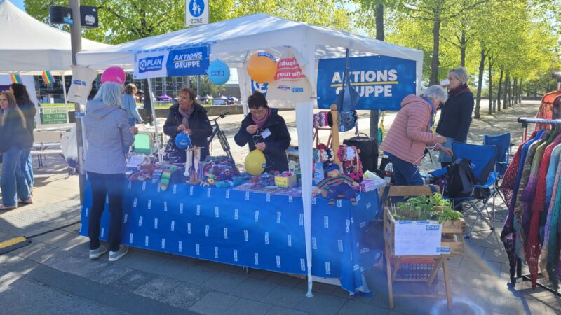Ein Marktstand im Freien mit einem weißen Vordach, auf dem „Aktions Gruppe“ steht. Mehrere Menschen stöbern in den ausgestellten bunten handgefertigten Waren und kleinen Pflanzen. Im Hintergrund sind Bäume, ein Kleiderständer und weitere Stände zu sehen.