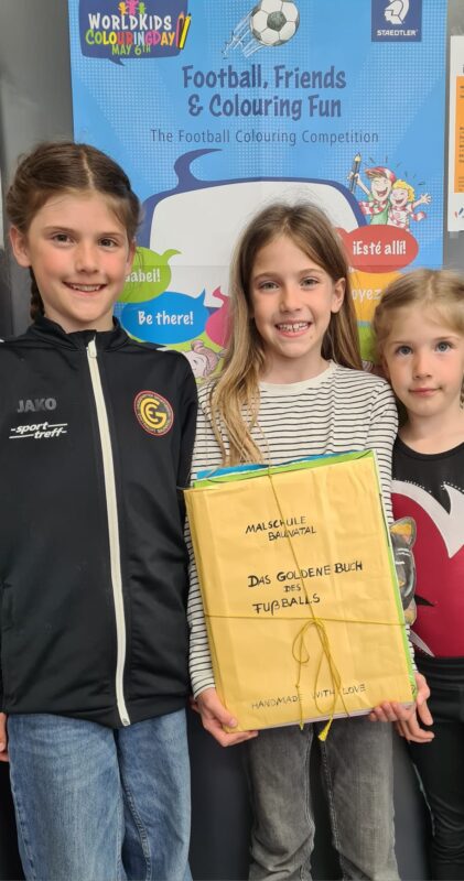Drei lächelnde Kinder stehen gemeinsam vor einem bunten Poster mit Fußballmotiven. Das mittlere Kind hält ein großes gelbes Buch mit der handgeschriebenen Aufschrift „Das goldene Buch des Fußballs“ in der Hand, das mit einer Schnur umwickelt ist.