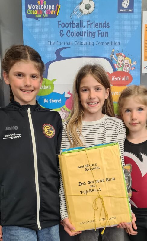 Drei lächelnde Kinder stehen gemeinsam vor einem bunten Poster mit Fußballmotiven. Das mittlere Kind hält ein großes gelbes Buch mit der handgeschriebenen Aufschrift „Das goldene Buch des Fußballs“ in der Hand, das mit einer Schnur umwickelt ist.